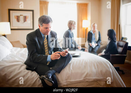 Businessman sits on the edge of a bed in a hotel room during a business trip and texts on his cell phone while his three female colleagues work together in the background. Stock Photo