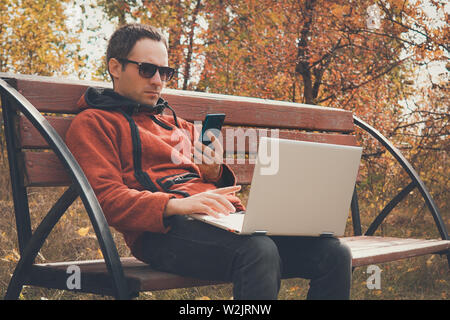 Young freelance man using mobile phone and laptop pc.Teenager student talking on smartphone in urban park. guy with wireless computer outdoor Stock Photo