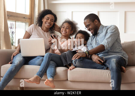 Cheerful african parents and kids laugh using devices on sofa Stock Photo