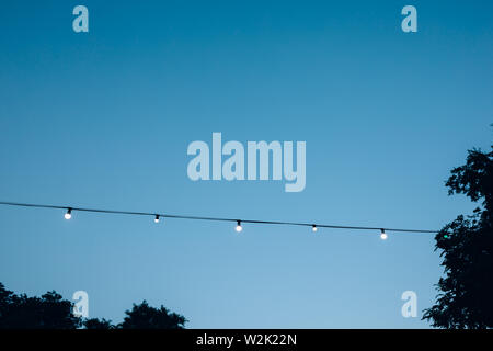 Decorative outdoor electric garland with light bulbs hanging on trees at summer evening. Stock Photo
