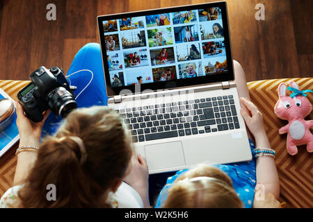 modern mother and child sitting on sofa in the modern living room viewing image library while transferring photos from DSLR photo camera on a laptop. Stock Photo