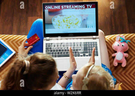 smiling young mother and child with credit card using streaming tv on a laptop while sitting on sofa in the modern living room. Stock Photo