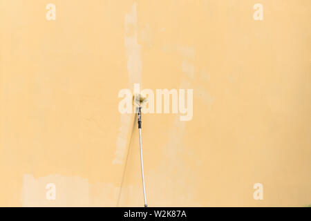 Painter paints building exterior wall with a roller. Roller with long stick manually painting building with yellow paint