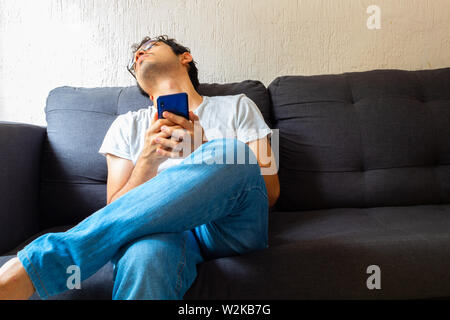 Young Hispanic man with clear brown skin wearing glasses using a bored smartphone while on a couch Stock Photo