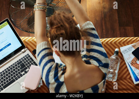 Upper view of trendy woman sitting on sofa at modern home in sunny hot summer day enjoying freshness in the front of working fan while suffering from Stock Photo