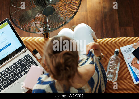 Upper view of young woman sitting on sofa in the modern living room in sunny hot summer day cooling down using metallic floor standing fan while suffe Stock Photo