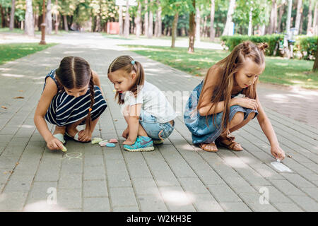 Three cute little girls sitting and drawing with chalk on asphalt in park in summer day Stock Photo