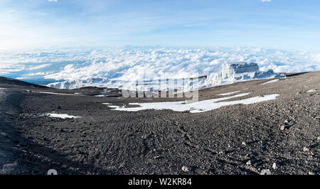 Footprints lead off through volcanic ash and dust towards a glacier sitting above the clouds on the summit of Mount Kilimanjaro, Tanzania. Stock Photo