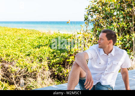 Young man on beach dunes in sunny day in Seaside, Florida panhandle town village looking at ocean leaning on railing bench in white shirt and jeans sh Stock Photo