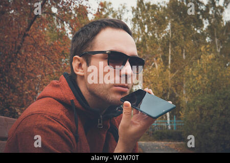 Young handsome man standing in park in summer, using a cell phone to listen to recorded voice message or audio. guy speaks into the microphone to Stock Photo