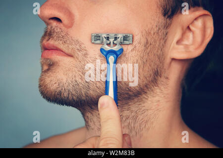 view of a young bearded man with a brutal razor. face on the blue background. big rough hairy bristles on the skin, side view close up Stock Photo