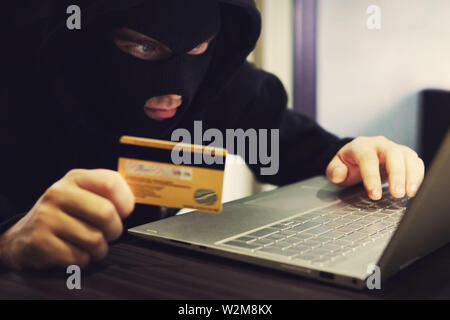 Man in robber mask and hood misappropriates personal bank data. Cyber fraudster attacks online banking system. Hacker makes financial fraud via Stock Photo