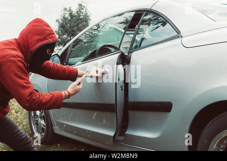 Car thief in hoodie managed to unlock the car using the smartphone and car link application. Man with masked face got the access to the vehicle cabin Stock Photo