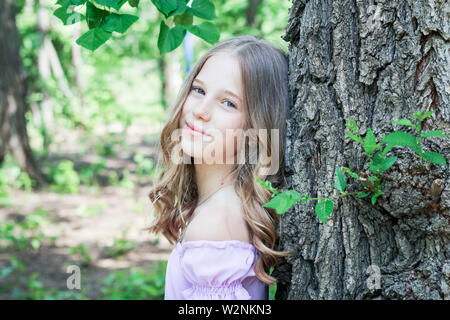 A young blonde girl in a flowered dress leaned back against a tree trunk in the park. Portrait of a young girl Stock Photo