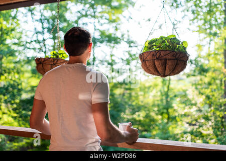 Hanging potted plants with violet flower leaves in spring with man standing on porch of house in morning wooden cabin cottage Stock Photo