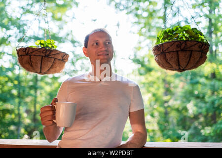 Hanging potted plants in spring with man standing on porch of house in morning wooden cabin cottage drinking coffee Stock Photo