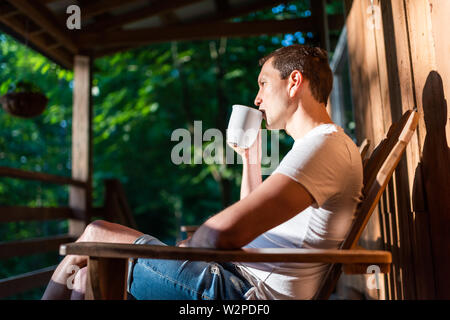 Man sitting relaxing on rocking chair on porch of house in morning wooden cabin cottage drinking coffee from cup Stock Photo