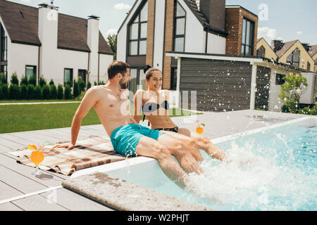 Happy couple sitting by the poolside in the backyard of their house. Man and woman sitting near water pool and funny splashing water their legs Stock Photo