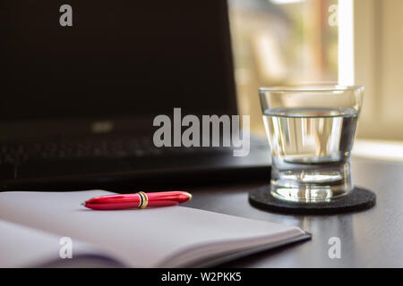 Home office in the morning with sunlight from window. Close up Glass of water, Laptop, Notebook with red pen on office desk. Office work concept Stock Photo