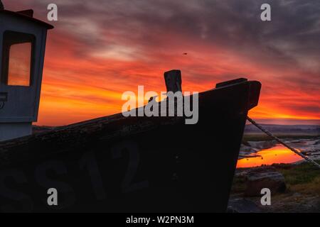 Small tugboat silhouetted against a fiery sky at sunrise, on the river bank Stock Photo