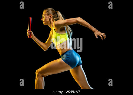 Professional female relay racer training isolated on black studio background in neon light. Woman in sportsuit practicing in running. Healthy lifestyle, sport, workout, motion, action concept. Stock Photo