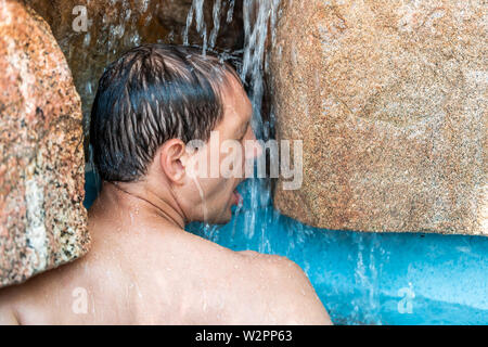 Young man swimming under waterfall in Japanese spa by stone pool colorful blue water in Japan onsen Stock Photo