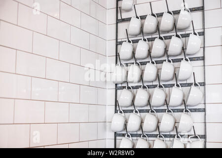 Close up of white cappuccino mugs hanging on a metal rack on white tiled wall. Stock Photo