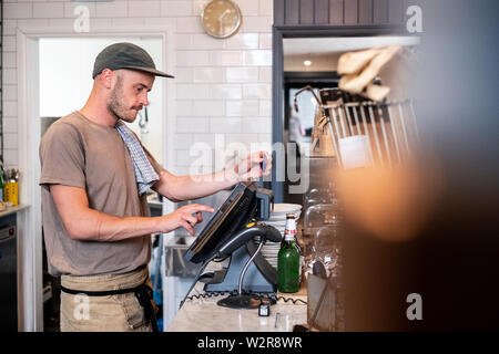 Bearded man wearing baseball cap standing at a till in a restaurant. Stock Photo