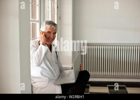 Doctor sitting in corridor, on the phone and using laptop. Stock Photo