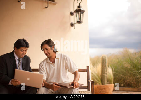 Two mid-adult men looking at a laptop while seated next to each other on a porch. Stock Photo