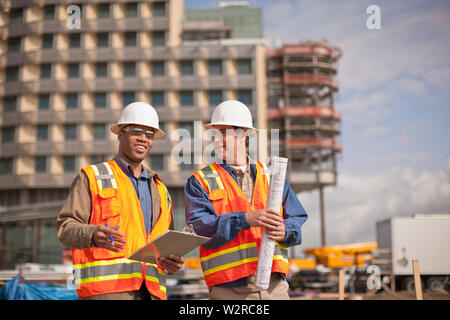 Two engineers in high visibility vests and hardhats discuss building plans on a construction site. Stock Photo
