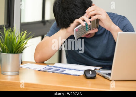 Asian man stressed while calculate the credit card debt with a calculator and working on laptop at home, Sad and dramatic,  Debt stress and mental hea Stock Photo