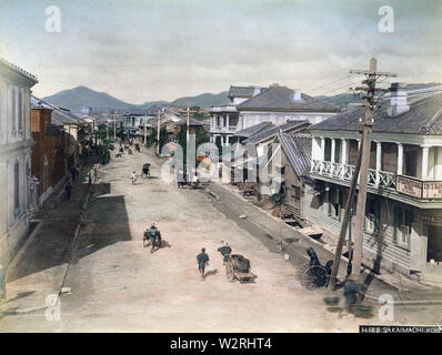 [ 1890s Japan - Main Street in Kobe ] —   A rickshaw puller rests in his carriage, while vendors go about their business, on Sakaemachi in Kobe, Hyogo Prefecture. The building on the right corner is the International Hotel.  19th century vintage albumen photograph. Stock Photo