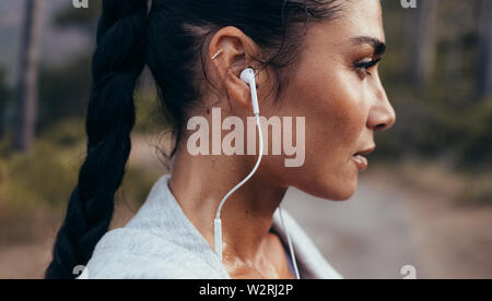 Side view of confident young woman with earphones standing outdoors after a morning run. Female athlete taking a rest after outdoor workout. Stock Photo
