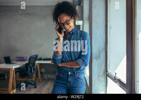 Positive young woman in casuals talking on mobile phone. Smiling female entrepreneur standing by a window in office and talking over cell phone. Stock Photo
