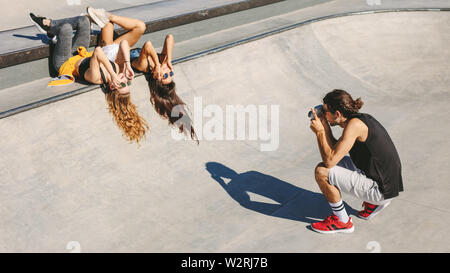 Two girls lying at skate bowl with a male friends taking their photograph. Photographer taking pictures of stylish women at skate park. Stock Photo