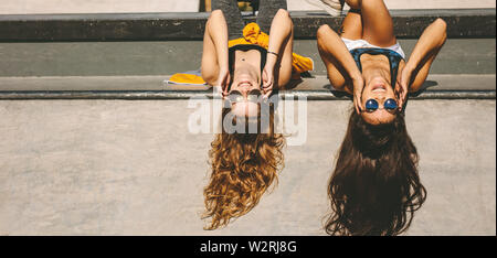 Two stylish girls lying down at skate park with sunglasses. Female friends having fun at skate park on a summer day. Stock Photo