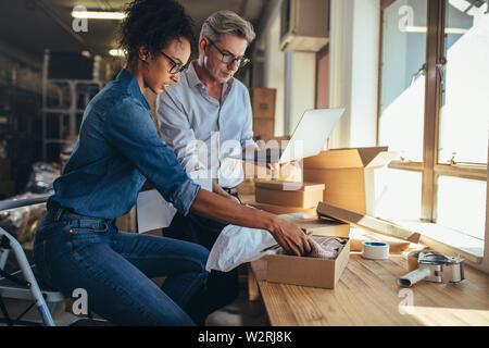 Online store partners preparing a parcel for shipping. Woman packing the product in box with man working on laptop. Stock Photo