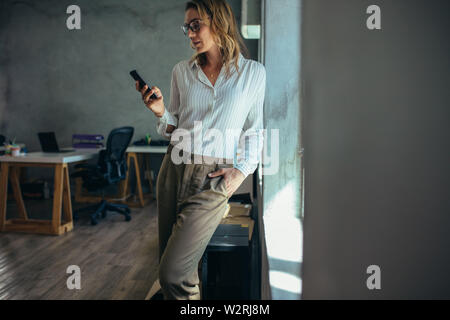 Businesswoman standing in office and using her smart phone. Female entrepreneur looking at her mobile phone. Stock Photo