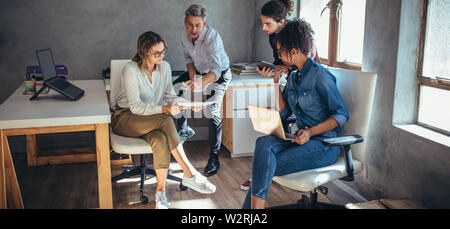 Diverse group of business people working together at a small office. Woman showing her digital tablet and discussing new business plan with coworkers. Stock Photo