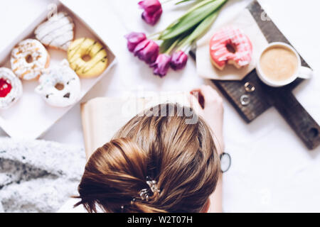 Girl reading a book and has brealfast in bed. Tray with strawberry pink donut with coffee and flowers. Background blurred. Top view. Stock Photo