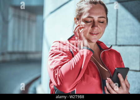 My favorite playlist. Happy plus size woman in headphones holding smart phone and listening music with closed eyes while standing against grey wall outdoors