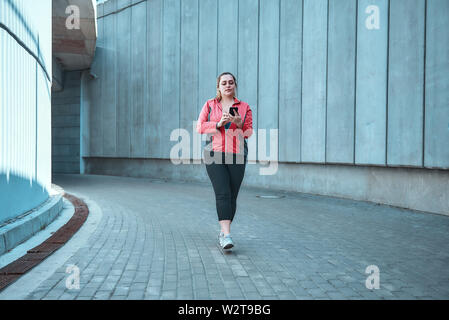 Ready for workout. Young plus size woman on sports clothes holding smart phone while standing outdoors. Digital technologies. Sport concept Stock Photo