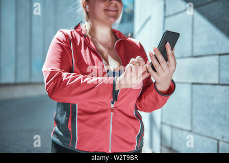 My favourite song Happy plus size woman in sports clothes and headphones touching her smart phone while standing outdoors. Digital technologies. Sport concept Stock Photo
