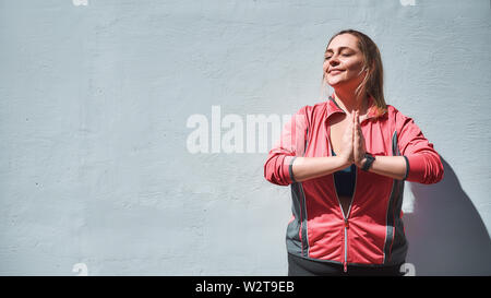 Life balance. Calm and pretty plus size woman in sporty clothes doing yoga while standing against wall with closed eyes. Yoga concept. Weight losing. Plump women Stock Photo