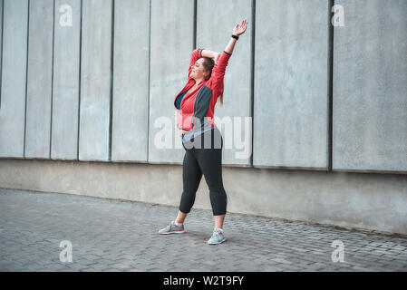 Beautiful plus size woman in sport clothes doing sport outdoors and smiling while standing against gray wall. Healthy life. Sport concept. Weight losing. Stock Photo