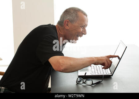 A man sits laughing loudly with open mouth as he points to the screen of a notebook computer. He reads or sees someting very funny on the screen. - Im Stock Photo