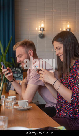 two friends sitting together in a cafe, using smart-phones. sitting together, yet each is using at their phone. Stock Photo