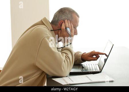 Casual dressed man pointing at computer screen and holding a pair of lasses in right hand. Papers lying He is looking a bit worried .  - Image Stock Photo