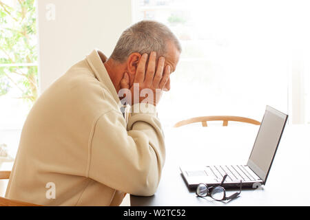 Casual dressed man sitting by a laptop computer is hiding his head in despair. A pair of glasses lying on a pile of papers next to the laptop. - Image Stock Photo
